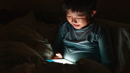 One cute Asian boy is enjoy watching the electronic tablet on the bed during bed time at night alone.Kid and the gadget using at home concept.