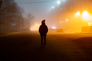 The dark silhouette of a human stands in the fog against the background of the lights of the houses...
