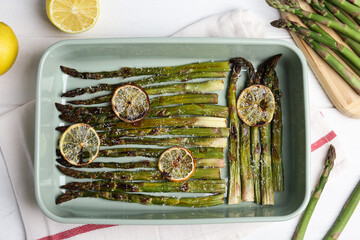 Oven baked asparagus with lemon slices in ceramic dish on white table, flat lay
