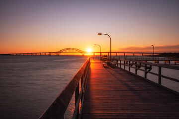Wooden fishing pier leading towards an arched bridge, as the sunset dips below the horizon. Captree...
