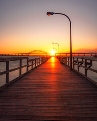 Wooden fishing pier leading towards an arched bridge, as the sunset dips below the horizon. Captree State Park - Long Island New York