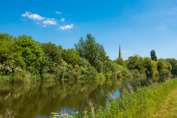 Obraz premium Lush riverbank seen during early summer near an inland waterway in Britain. A distant church spire is located against a near clear blue sky.