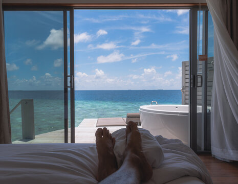 A legs man on the white bed with luxury beautiful room design on resort, view from the room on clear blue sea, summer vacation in Maldive.