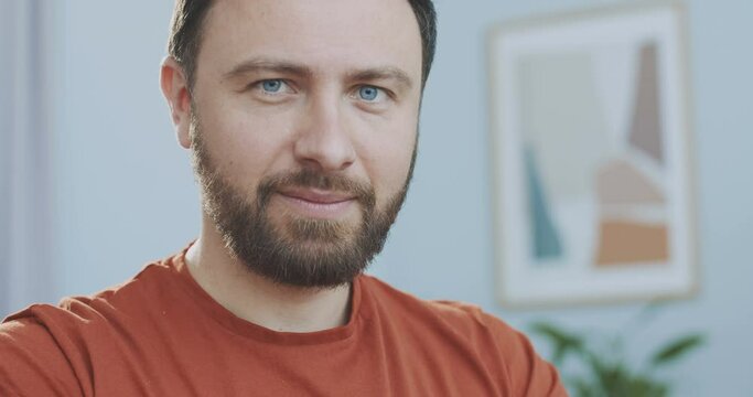 Close up portrait of the caucasian good looking young man with blue eyes and red t-shirt with a beard smiling to the camera sitting at home office. Concept of freelance, home office
