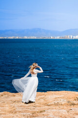 Bride with wreath in white wedding dress on the rock beach against beautifull blue sea background. Blond bride touching her hair on the beach. Concept of wedding and bridal morning. wedding Mallorca,