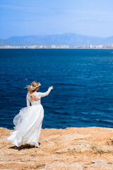 Obraz premium Bride with wreath in white wedding dress on the rock beach against beautifull blue sea background. Blond bride pointing at sea on the beach. Concept of wedding and bridal morning. wedding Mallorca,