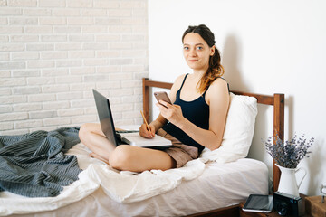 young European woman using a laptop works remotely from home, in her bed, talking on smartphone with a client, while the virus is quarantined.Cozy workplace, e-learning concept.freelancer