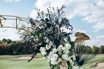 A wedding arch stands on a bare field and is decorated with tropical plants, golden flowers and white roses. Wedding ceremony in the open air.