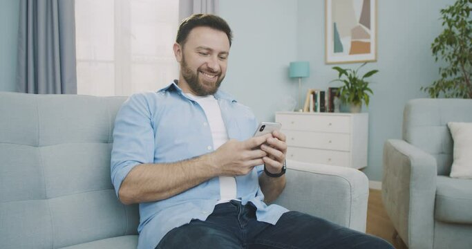 Middle plan of cheerful middle aged man sitting on the sofa using phone smile in the modern apartment texting message scrolling tapping. Concept of technology, isolated, lifestyle