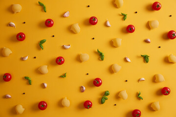 Uncooked pasta, red tomatoes, basil leaves and peppercorns on yellow background. Ingredients for preparing delicious pasta. Traditional Italian food or cuisine. Products for cooking. Top view