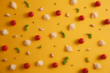Flat top view of pasta shells made of durum wheat, red cherry tomatoes, basil and garlic for preparing Italian dish. Vegetables and spices on yellow background. Cooking ingredients. Detox food
