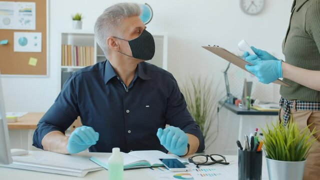 Man in mask is busy with computer work while female employee is taking his temperature with infrared thermometer concerned with safety in workplace.