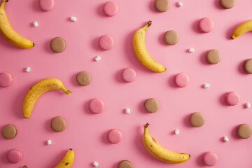 Assortment of sweet delicious colorful french macaroons, white marshmallow and bananas on pink studio background. Unhealthy high calories cookies and healthy tropical fruit. Idea for your dessert