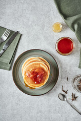 American-style pancakes with apple jam in muted green plate on grey background. Fork and knife on a napkin, jars with honey, tea spoon and some tea on the table