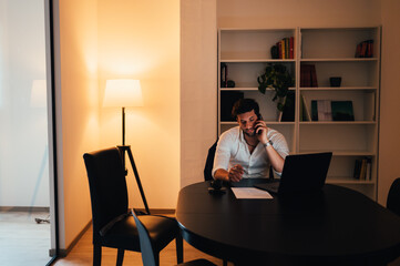 Young businessman talking on the phone and using a laptop computer at his desk in the office or home late into the night