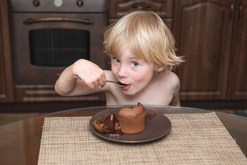 .little boy with blond hair sits at a table in the kitchen and eats with a spoon a chocolate dessert and look at the camera