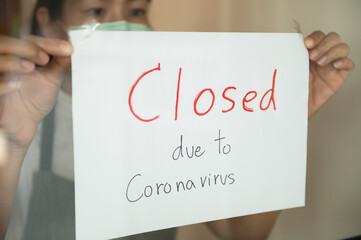 Young Asian business owner coffee shop wearing face surgical mask announcing shop be closed by pasting closed sign at shop entrance due to business crisis from coronavirus outbreak problem.