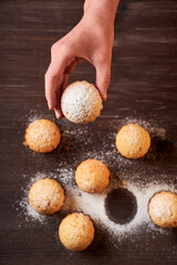 Freshly baked muffins with some icing sugar on dark brown wooden table, woman’s hand holding one muffin above the table, top view vertical shot