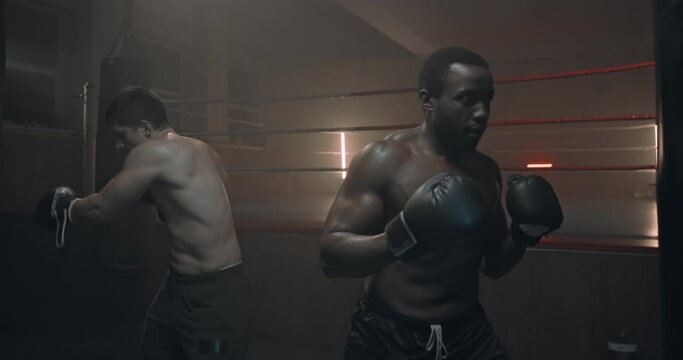 Multiethnic young male shirtless kickboxers punching bag in dark ring at training. Caucasian and African American sweaty men exercising and kickboxing in darkness. Mixed-races sportsmen in gloves.