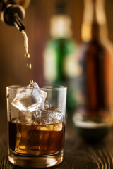 In the bar, whiskey is poured into a glass with ice, in the background there are bottles on a wooden table of the bar counter, shallow depth of field, selective focus. The concept of alcoholic drinks