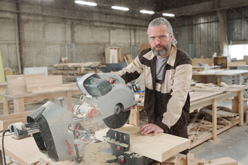 Confident aged professional in eyeglasses and workwear cutting wooden board