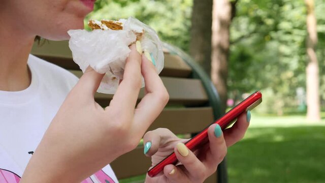 Female in white T-shirt reads message on red smartphone. Woman eats piece of delicious cake. Summer park background. Break time concept