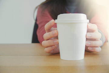 Closed up image of a woman sitting with hands holding a white paper coffee cup. White background and wood table.