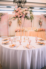 Banquet, restaurant. Table setting. White plates with a gold rim, golden cutlery, glasses. Peach-colored napkin with a golden ring. Peach pink floral arrangements. Tall tapers in gold candle holders