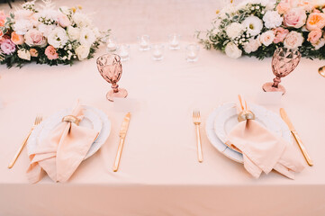 Banquet, restaurant. Table setting. White plates with a gold rim, golden cutlery, glasses. Peach-colored napkin with a golden ring. Peach pink floral arrangements
