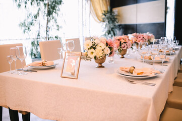 Banquet, restaurant. Table setting. White plates with a gold rim, golden cutlery, glasses. Peach-colored napkin with a golden ring. Peach pink floral arrangements