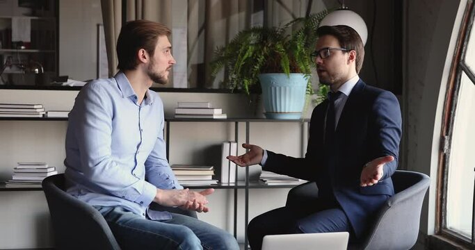 Two male executive business partners working together, talking, discussing corporate strategy at meeting sitting in chairs. Serious professional businessmen having conversation in modern office.