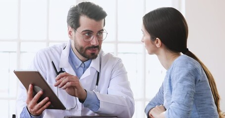 Confident young male general practitioner in white uniform showing lab test results on digital computer tablet to focused female patient at clinic meeting, medical worker using tech healthcare apps.