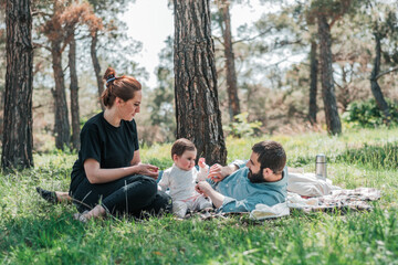 Happy young family with small 8 month daughter having picnic in forest. Cute, Happy, Funny family concept. Father and Mother are playing with daughter and smiling.