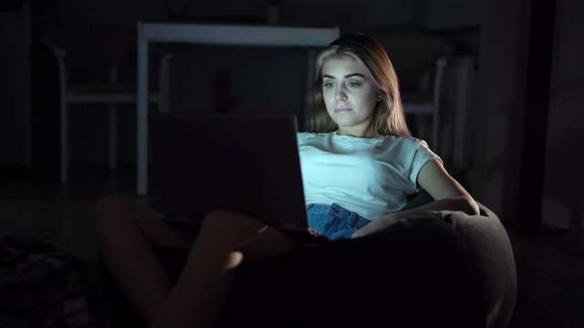 Beautiful girl sitting in a chair and watching a movie on a laptop. She is alone in a dark room. Online cinema. Watching videos at home.