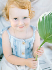 Vertical portrait of ginger red heared blue eyed cute kid child baby girl sitting outdoor smiling at camera happy holding green leaf on hands learning basic biology respect and protect nature