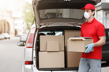 Delivery man with protective mask and gloves holds box near open car with many packages