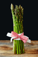 Bunch of asparagus stems tied up with two ribbons - red and white. Standing vertically on a rustic wooden table. Black background. Copy space, space for text. Healthy food