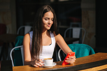 Attractive young woman using phone in cafe on terrace. Social distancing. Beautiful brunette girl chatting and drink coffee