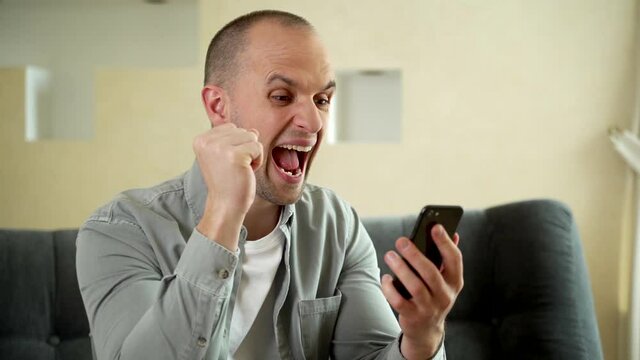Man using smartphone and winning big money on sports betting. Guy sitting on sofa and rejoicing victoryb while money falling from above