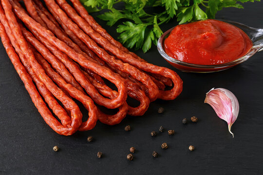 Bunch of thin dry smoked sausage kabanos or cabanossi and tomato ketchup on a black stone serving board. Traditional polish meat delicacy.