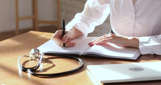Close up focus on female doctor hands writing treatment prescription notes. Professional male general practitioner in white uniform making medical records in paper notebook, working at clinic office.