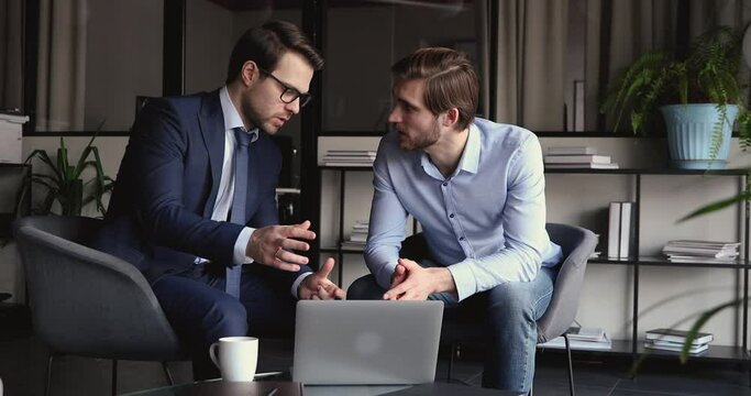 Serious male executive wearing suit consulting business partner or client showing corporate project online presentation on computer at meeting. Two businessmen looking at laptop and talking in office.