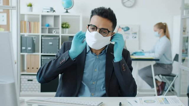 Portrait of Arab man putting on facial medical mask in office during epidemic sitting at desk and looking at camera. Woman is working in background.