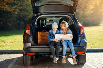 Agreeable boy and girl are looking at the road map while sitting in the auto's trunk and discussing the move direction. Family vacation trip by car.