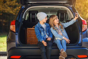 Front view of handsome 10-aged boy in hat and pretty 12-aged girl with blond long hair which taking photos on her phone,sitting in the car's trunk