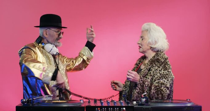Glamorous vintage stylish grandmother and gradfather dancing and having fun at DJ mixer on pink wall background. Couple of old retro male and female djs playin music on equipment.
