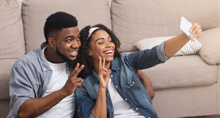 Happy african couple posing for selfie at home, using camera on smartphone