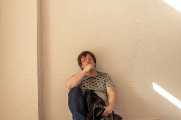 A man with glasses wistfully sits leaning on a wall. He's wearing jeans and a t-shirt. In his hand...