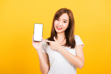 Naklejka premium Asian happy portrait beautiful cute young woman smile standing wear t-shirt making finger pointing on smartphone blank screen looking to camera isolated, studio shot yellow background with copy space