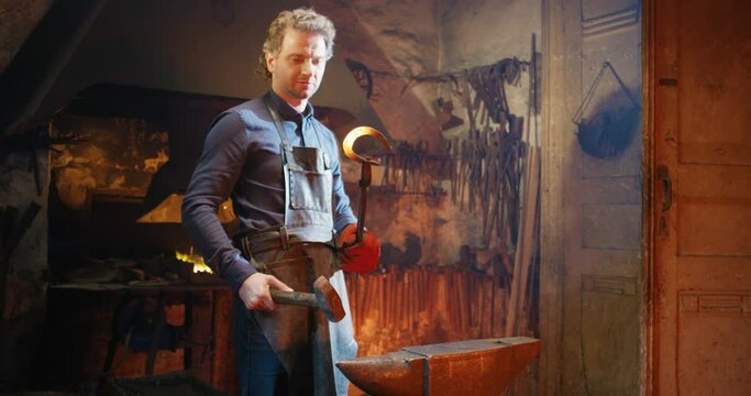 Handsome Caucasian blacksmith working in antique smithy and forging glowing horseshoe with hammer. Male young forger hard work in heat of metal manufactory.